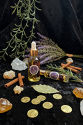 Two glass dropper bottles of 'The Magic' essential oil by House of the Crescent, one standing upright and one lying horizontally, styled with lavender, rosemary, a crystal, and black fabric on a dark stone surface.